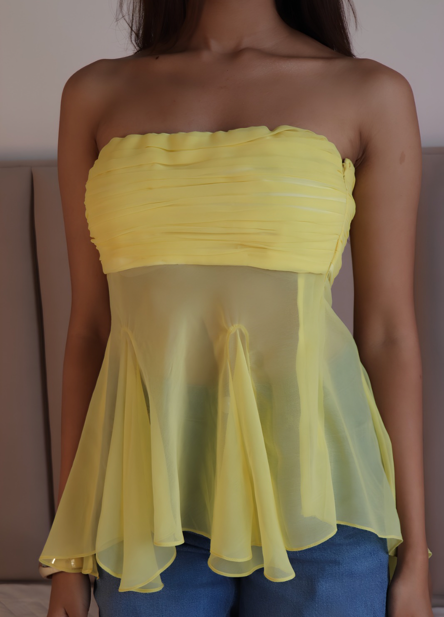 A woman wearing a yellow strapless top with a ruched bust and a sheer mesh peplum overlay, paired with blue jeans.